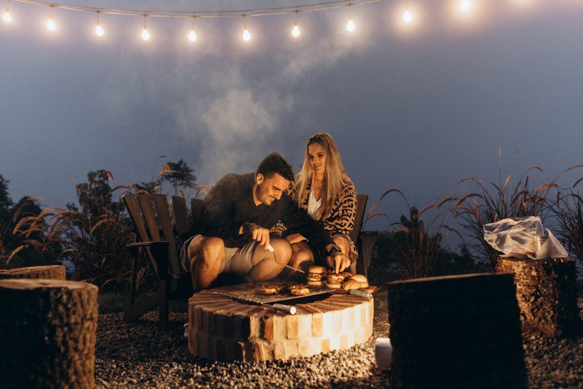 Couple enjoying a cozy barbecue by a fire with glowing string lights, creating a warm, romantic evening.