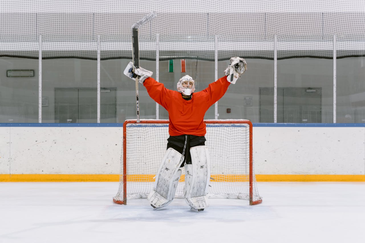 Ice hockey goalie in red jersey celebrating with raised arms in the rink's goal area.