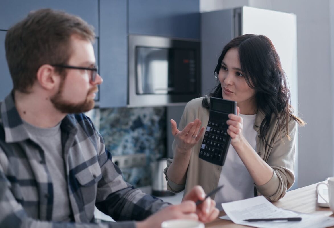 A couple sitting in a kitchen discussing financial matters using a calculator and documents.